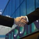 Two people shaking hands as they secure a new deal in front of a building with a large Bitcoin sign on its front.