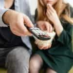 A man and a woman sitting on a sofa, pointing a TV remote and eating popcorn.