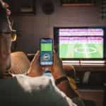 A man sits on a sofa watching a soccer game and using a smart phone to place a bet.