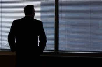 The dark silhouette of a man in a suit looking through the thin blinds of the windows in his office.