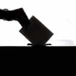 A silhouette of a person’s hand slipping an envelope into a ballot box during an election.