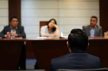 From behind, a man sits in front of a panel of three observers in a courtroom.