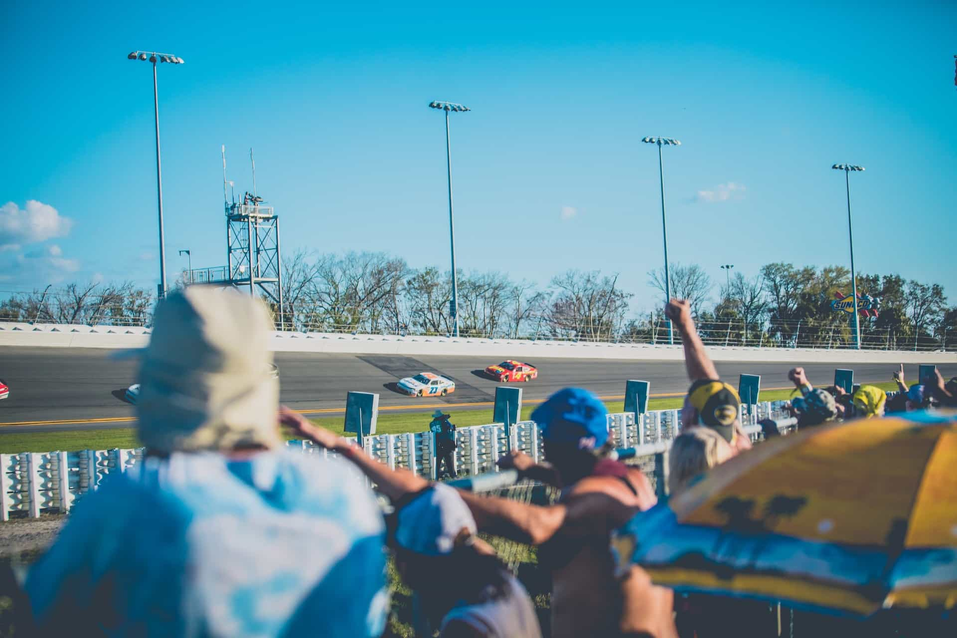 The curved bend of an outdoor NASCAR track during a race, with spectators cheering the passing vehicles.